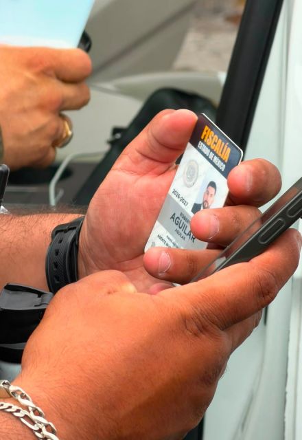 Mano sostiene una credencial de la Fiscalía del Estado de México junto a un teléfono celular durante una intervención policial.