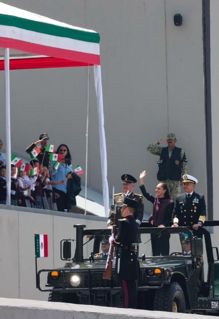 La Presidenta Electa Claudia Sheinbaum saludando y sonriendo desde un vehículo militar abierto durante el desfile conmemorativo en el Heroico Colegio Militar de Tlalpan, acompañada de mandos de la SEDENA y SEMAR.