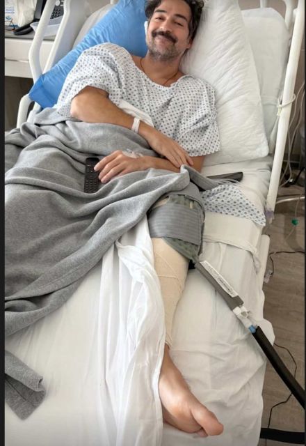 Alfonso Herrera sonríe en una cama de hospital, con bata médica, pierna vendada y equipo médico tras cirugía de rodilla.