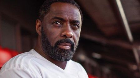 Idris Elba con playera blanca sentado en bancas rojas