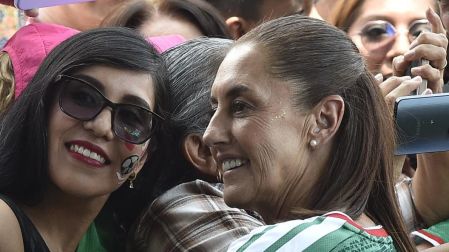 La presidenta Claudia Sheinbaum sonriendo y posando para una selfie detrás de una valla metálica con un grupo de aficionados de la Selección Mexicana.