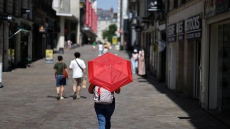 Una persona caminando por una calle peatonal de tiendas en Nantes, sosteniendo un paraguas rojo abierto como parasol para protegerse del sol abrasador.