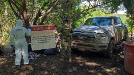 Elemento de la Marina con traje de protección hazmat y personal táctico sosteniendo una lona de erradicación frente a una camioneta RAM asegurada.