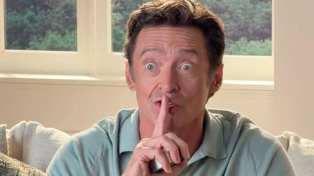 Hugh Jackman con camisa polo azul claro, sentado en un sofá y colocando su dedo índice sobre los labios.