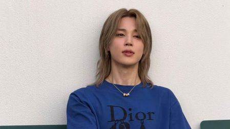 Jimin de BTS sentado en una banca verde, con cabello rubio, camiseta azul Dior y pantalón de mezclilla.