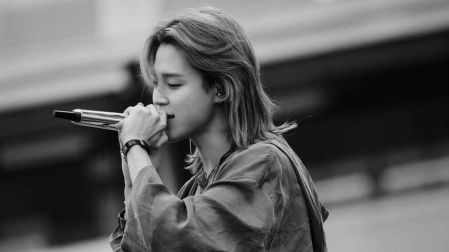 Jimin de BTS cantando con un micrófono plateado, cabello largo y sudadera holgada durante una presentación en blanco y negro.
