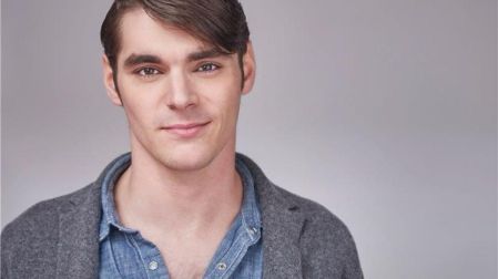 RJ Mitte, actor de Breaking Bad, con camisa azul de mezclilla y saco gris, posando frente a un fondo gris claro.
