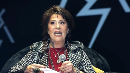 Alejandra Guzmán con chamarra negra de estoperoles y vestido rojo, sentada en un sillón mientras sostiene un micrófono.