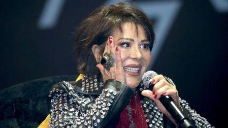 Alejandra Guzmán con chamarra negra de estoperoles, sonríe mientras sostiene un micrófono y se lleva una mano al rostro.