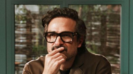 Alfonso Herrera con lentes negros y saco café, sentado frente a una ventana con una mano cerca de la boca.