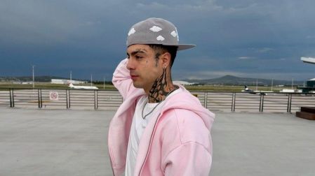 Naim Darrechi posa de perfil con sudadera rosa, gorra gris y tatuajes en el cuello, frente a la pista de un aeropuerto.