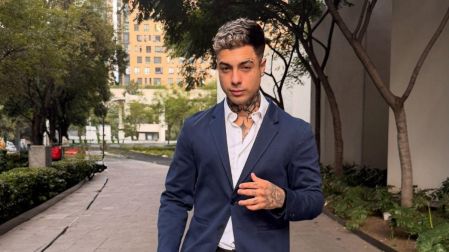 Naim Darrechi posa con saco azul marino, camisa blanca y pantalón negro en una calle arbolada, mostrando tatuajes en cuello y manos.