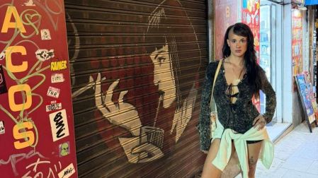 Gala Montes posa en una calle con vestido oscuro, chamarra atada a la cintura y bolso, frente a una cortina metálica con grafiti.