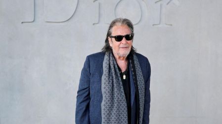 Al Pacino posa con lentes oscuros, saco azul marino y bufanda gris frente a una pared con el logotipo de Dior.