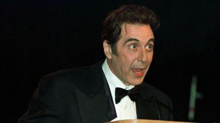 Al Pacino joven con esmoquin negro, camisa blanca y moño, hablando frente a un micrófono detrás de un podio de madera.