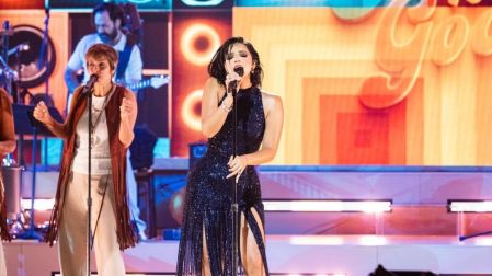 Ángela Aguilar canta con vestido azul brillante y micrófono en el escenario, acompañada de otra cantante y músicos.