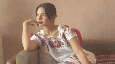 Ángela Aguilar sentada en un sofá gris, con vestido blanco de flores bordadas, cabello recogido y sandalias azules.