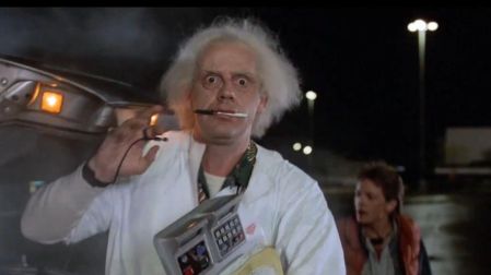 Doc Brown sostiene un dispositivo junto al DeLorean mientras Marty McFly aparece detrás en una escena nocturna de Volver al futuro.
