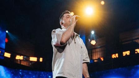 Josi Cuen canta con micrófono y camisa gris sobre un escenario iluminado durante una presentación musical.