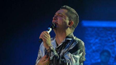 Josi Cuen canta con un micrófono y viste una camisa estampada durante un concierto con iluminación azul.
