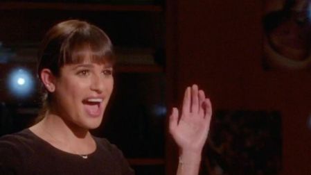 Lea Michele como Rachel Berry en Glee, con flequillo, blusa negra y una mano levantada mientras canta.