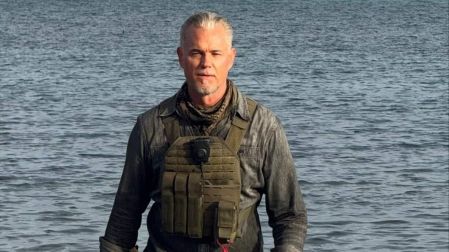 Eric Dane frente al mar, con cabello canoso, camisa gris oscura, chaleco táctico y pantalones negros.