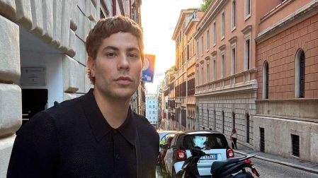 Christian Chávez viste camisa negra y posa en una calle empedrada con edificios, automóviles y una motocicleta al fondo.