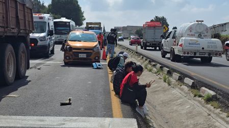 Personas sentadas en el camellón central de la carretera Apizaco-Tlaxcala junto a un auto naranja chocado por la parte frontal y una ambulancia, esperando atención tras choque múltiple.