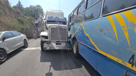 Tráiler de carga color blanco impactado directamente contra el costado de un microbús de pasajeros de la empresa ATAH en la carretera Apizaco-Tlaxcala. Al fondo se observa una camioneta volcada.