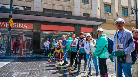 Grupo de trabajadores del Gobierno de la CDMX y ciudadanos sosteniendo escobas durante el lavado de una calle empedrada en el Centro Histórico, frente a comercios locales.