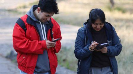 Un hombre joven con chamarra roja y una mujer joven revisando sonrientes sus teléfonos celulares al aire libre, representando a los usuarios de telefonía móvil.