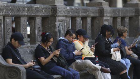 Fila de personas de diferentes edades sentadas en una banca de piedra en la calle, todas concentradas mirando las pantallas de sus teléfonos celulares.