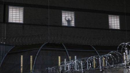 Silueta de una persona detrás de una ventana iluminada y enrejada en un centro de detención de ICE, rodeado de cercas con alambre de púas durante la noche.