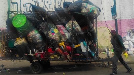 Hombre caminando junto a un remolque desbordado de bolsas de basura y plásticos reciclables en calles de CDMX.