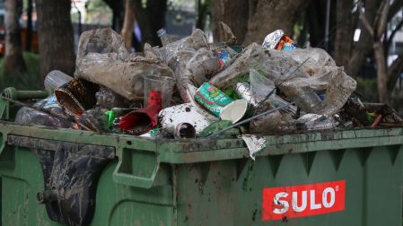 Bote de basura público color verde completamente desbordado con botellas de plástico, vasos y envolturas.