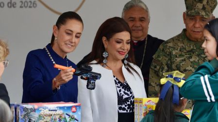 Claudia Sheinbaum sosteniendo y examinando una pistola de juguete negra con decoraciones frente a una niña, durante el intercambio por juguetes educativos y de destreza.