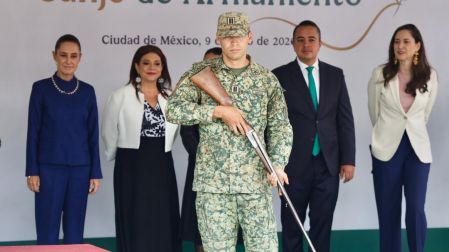 Un elemento militar de la Secretaría de la Defensa Nacional sosteniendo un arma de fuego larga durante el programa Canje de Armamento, con la Presidenta Claudia Sheinbaum observando en el fondo.