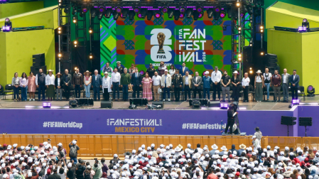 Vista elevada del gran escenario verde del FIFA Fan Festival 2026 en Ciudad de México, con el logo oficial y una fila de autoridades, frente a una multitud con gorras blancas.