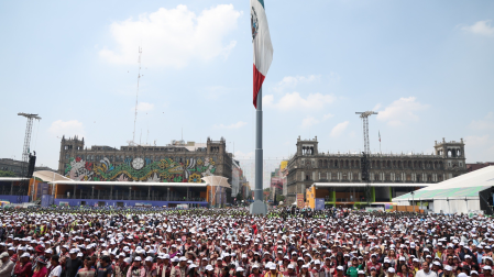 Vista panorámica de miles de servidores públicos de CDMX con gorras blancas llenando la plancha del Zócalo durante el Fan Fest del Mundial 2026, frente a edificios históricos decorados.