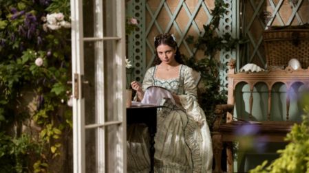 Joven Violet Ledger, personaje de La reina Carlota, sentada en un jardín mientras borda, con vestido de época color verde claro.