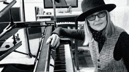 Diane Keaton con sombrero y lentes, sentada junto a un piano en una fotografía en blanco y negro.
