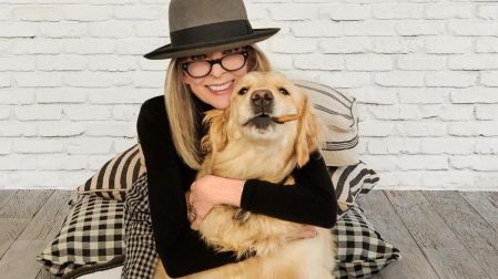 Diane Keaton con sombrero gris y lentes negros, abrazando a un perro golden retriever frente a una pared blanca.