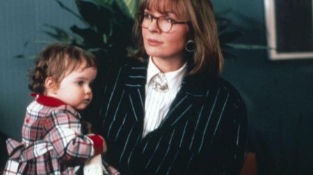 Diane Keaton con lentes y traje negro de rayas, sosteniendo a una niña pequeña vestida a cuadros.