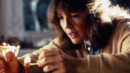 Diane Keaton con cabello castaño y suéter beige, encendiendo un fósforo junto a una vela roja en una escena cinematográfica.