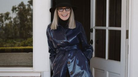 Diane Keaton con sombrero negro, lentes y abrigo azul brillante, sonriendo frente a una puerta blanca.