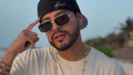 Eduin Caz, vocalista de Grupo Firme, con camiseta blanca, gorra negra, lentes oscuros y cadena dorada al aire libre.