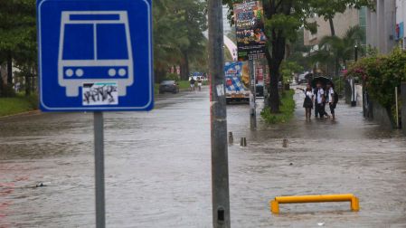 Grupo de jóvenes caminando con el agua por encima de los tobillos cerca de un letrero de parada de autobús en una avenida inundada de Acapulco.