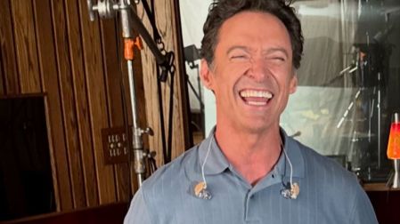 Hugh Jackman sonríe con los ojos cerrados, vestido con una camisa polo azul y audífonos de monitoreo alrededor del cuello.