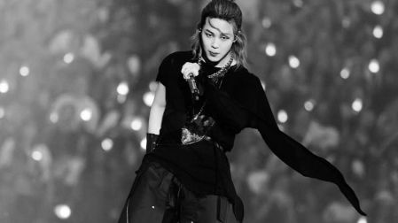 Jimin de BTS durante un concierto en blanco y negro, vestido de negro, con cabello recogido y micrófono en mano.