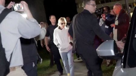Selma Blair con sudadera blanca, jeans y lentes oscuros, rodeada de policías y periodistas durante la noche en Nueva York.